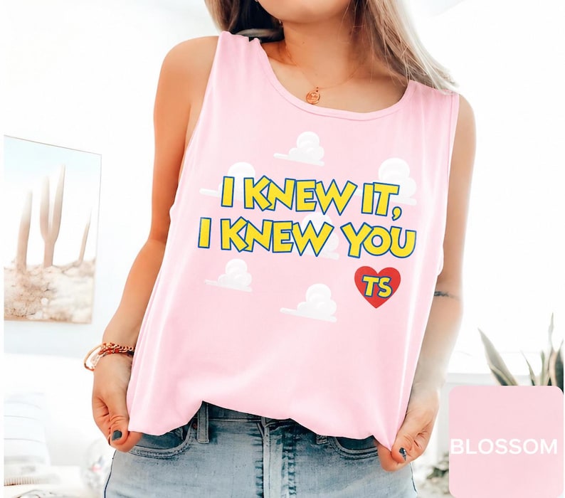 May include: A light pink sleeveless tank top featuring a graphic print of white clouds and the yellow text "I KNEW IT, I KNEW YOU" with a red heart containing the letters "TS" in yellow. The garment has a round neckline and a relaxed fit.