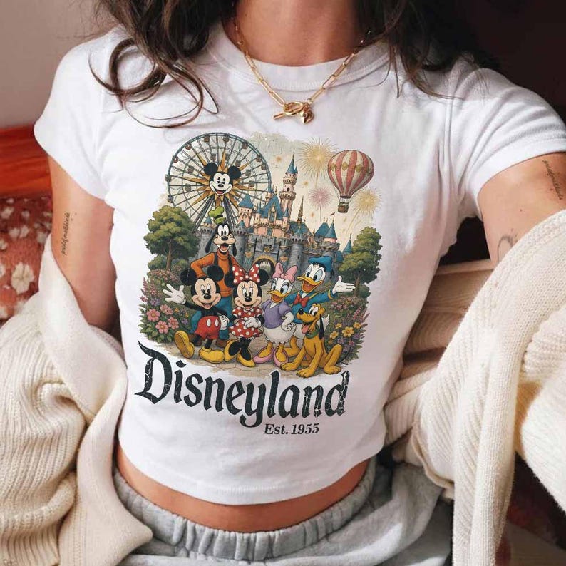 May include: A white short-sleeve t-shirt featuring a colorful graphic of Mickey Mouse, Minnie Mouse, Goofy, Donald Duck, Daisy Duck, and Pluto in front of a castle, a ferris wheel, and a hot air balloon. The text reads Disneyland Est. 1955.