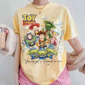 May include: Yellow t-shirt featuring a graphic of characters from the animated film "Toy Story." The design includes Woody, Buzz Lightyear, and the Little Green Men, with the text "Toy Story" and "You've got a Friend in me."