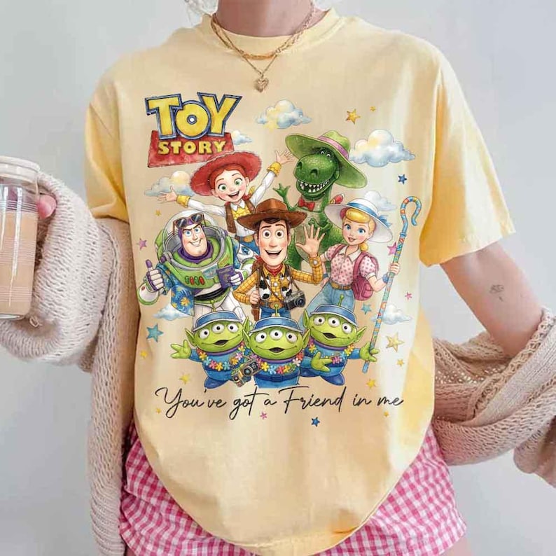May include: Yellow t-shirt featuring a graphic of characters from the animated film "Toy Story." The design includes Woody, Buzz Lightyear, and the Little Green Men, with the text "Toy Story" and "You've got a Friend in me."