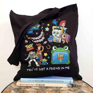 May include: A black tote bag featuring a colorful graphic print of Disney Pixar Toy Story characters including Woody, Buzz Lightyear, and Jessie. The design includes the text Disney Pixar, Toy Story 5, and the phrase You've Got A Friend In Me.