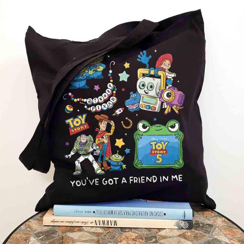 May include: A black tote bag featuring a colorful graphic print of Disney Pixar Toy Story characters including Woody, Buzz Lightyear, and Jessie. The design includes the text Disney Pixar, Toy Story 5, and the phrase You've Got A Friend In Me.