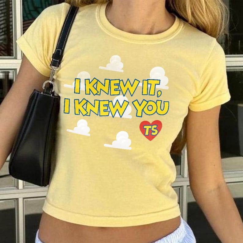 May include: A pale yellow short-sleeved t-shirt featuring a graphic print of white clouds and the text "I KNEW IT, I KNEW YOU" in blue letters with yellow outlines, accompanied by a red heart containing the letters "TS" in yellow.
