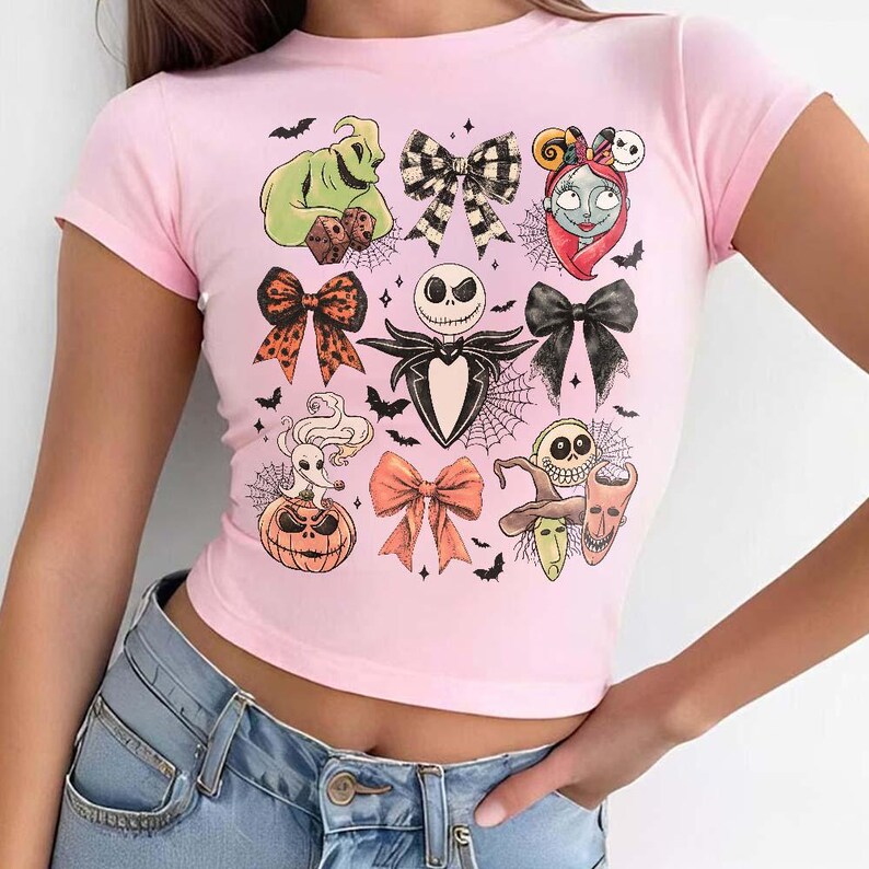 May include: A light pink crop top featuring various illustrations from The Nightmare Before Christmas. The design includes characters like Jack Skellington, Sally, and Oogie Boogie, along with bows, bats, and spiderwebs. The shirt has short sleeves.
