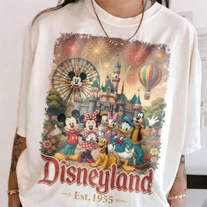 May include: A white t-shirt featuring a graphic print of Mickey Mouse, Minnie Mouse, Daisy Duck, Donald Duck, Pluto, and Goofy in front of a castle, a Ferris wheel, and fireworks. The text Disneyland Est. 1955 is printed in red at the bottom.