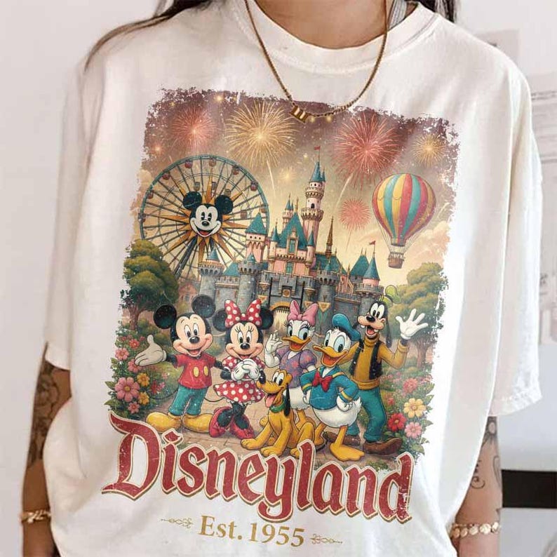 May include: A white t-shirt featuring a graphic print of Mickey Mouse, Minnie Mouse, Daisy Duck, Donald Duck, Pluto, and Goofy in front of a castle, a Ferris wheel, and fireworks. The text Disneyland Est. 1955 is printed in red at the bottom.