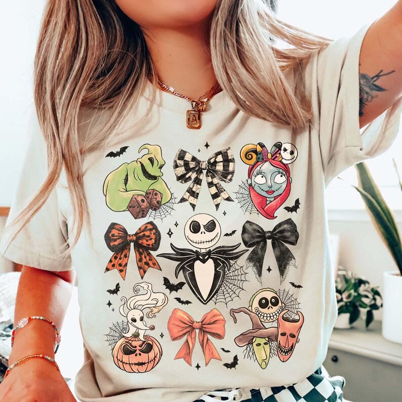 May include: Cream-coloured t-shirt featuring Halloween character illustrations. The design includes Jack Skellington, Sally, Oogie Boogie, and other characters, along with bows, bats, and spiderwebs. A spooky themed top.