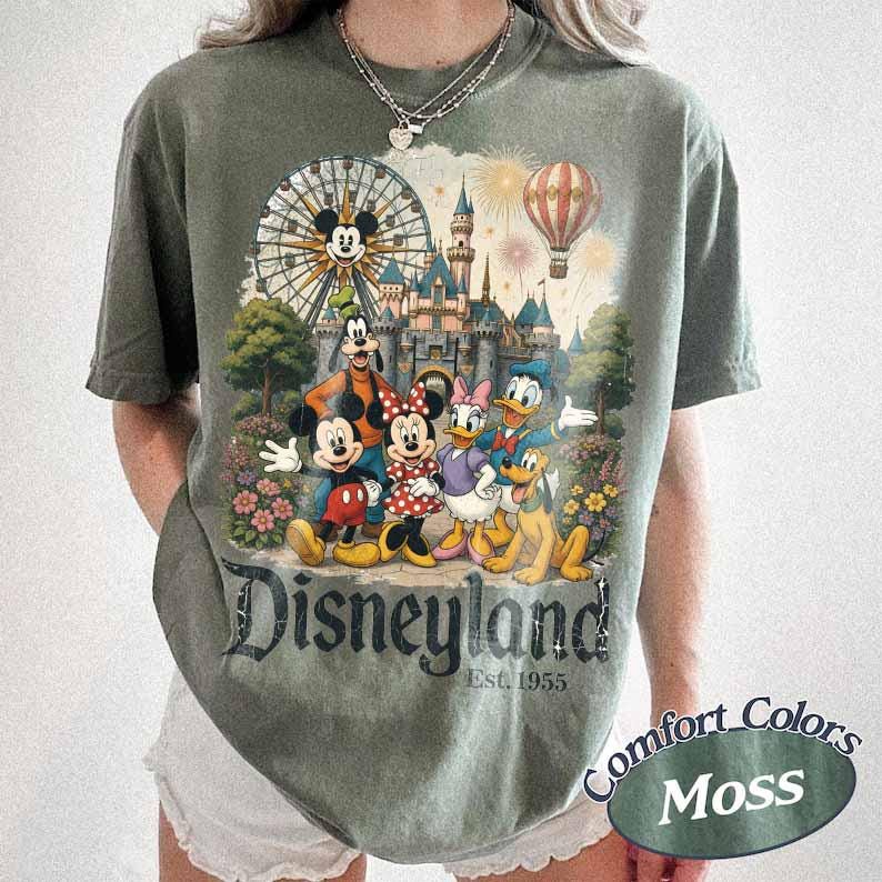 May include: A moss green short-sleeve t-shirt featuring a graphic print of Mickey Mouse, Minnie Mouse, Goofy, Donald Duck, and Pluto in front of a castle, a Ferris wheel, and a hot air balloon. The text reads Disneyland Est. 1955 in a dark font.
