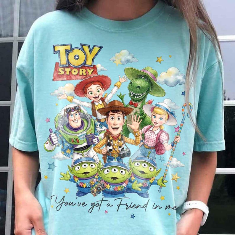 May include: A light blue t-shirt featuring a colorful graphic of characters from the animated film "Toy Story." The design includes Woody, Buzz Lightyear, Jessie, Rex, and the Little Green Men, with the text "Toy Story" and "You've got a Friend in me."