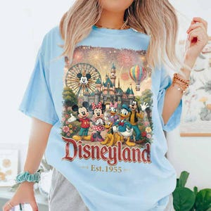 May include: A light blue t-shirt featuring a graphic print of Mickey Mouse, Minnie Mouse, Donald Duck, Goofy, and Pluto in front of a Disneyland castle, a Ferris wheel, and a hot air balloon. The text reads Disneyland Est. 1955 in red lettering.