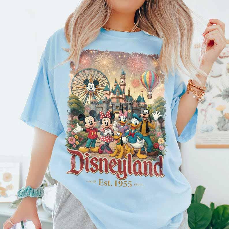 May include: A light blue t-shirt featuring a graphic print of Mickey Mouse, Minnie Mouse, Donald Duck, Goofy, and Pluto in front of a Disneyland castle, a Ferris wheel, and a hot air balloon. The text reads Disneyland Est. 1955 in red lettering.