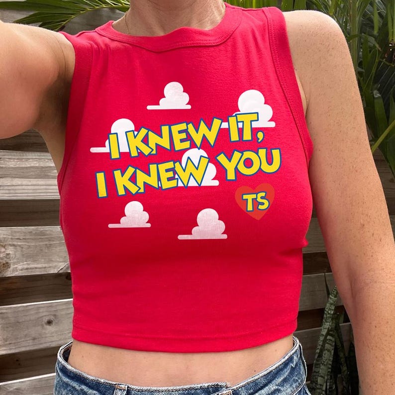 May include: A bright red sleeveless crop top featuring a graphic print of white clouds. The text reads I KNEW IT, I KNEW YOU in yellow letters with a blue outline, accompanied by a red heart shape containing the yellow letters TS.