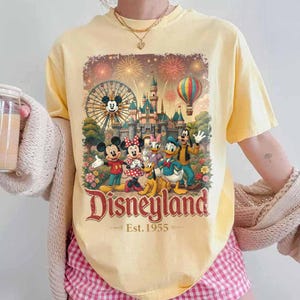 May include: A pale yellow short-sleeve t-shirt featuring a graphic print of Mickey Mouse, Minnie Mouse, Donald Duck, Goofy, and Pluto in front of a castle, a Ferris wheel, and a hot air balloon with the text Disneyland Est. 1955 in red lettering.