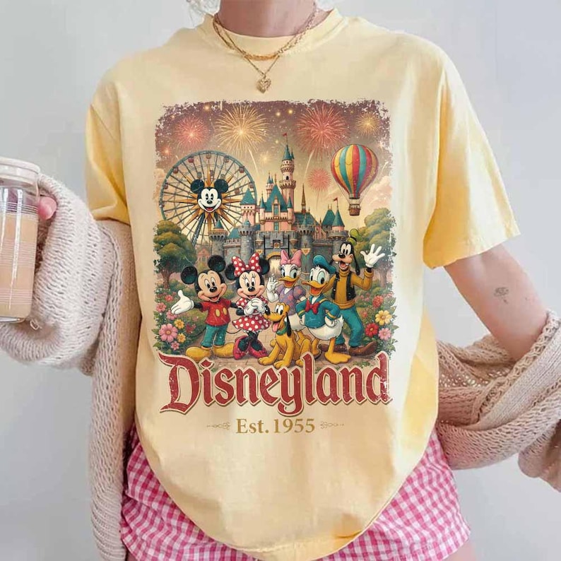 May include: A pale yellow short-sleeve t-shirt featuring a graphic print of Mickey Mouse, Minnie Mouse, Donald Duck, Goofy, and Pluto in front of a castle, a Ferris wheel, and a hot air balloon with the text Disneyland Est. 1955 in red lettering.