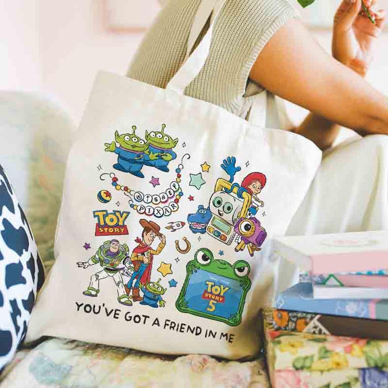 May include: A cream-colored canvas tote bag featuring a colorful Toy Story graphic print. The design includes characters like Woody, Buzz Lightyear, and aliens, along with the text Disney Pixar, Toy Story, Toy Story 5, and You've Got a Friend in Me.