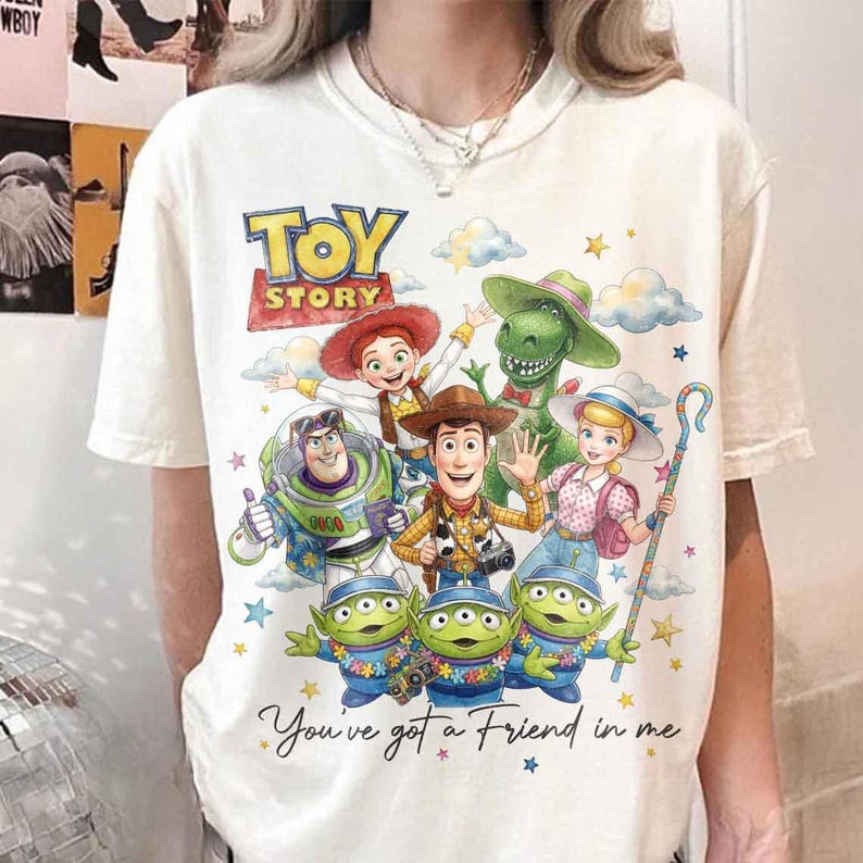 May include: Cream-colored t-shirt featuring a colorful graphic of Toy Story characters, including Woody, Buzz Lightyear, and the Little Green Men. The text "TOY STORY" is at the top, with "You've got a Friend in me" at the bottom.