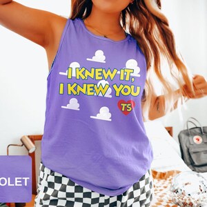 May include: A violet sleeveless tank top featuring a repeating white cloud pattern and yellow text that reads I KNEW IT, I KNEW YOU with a red heart containing the letters TS. The garment has a round neckline and a relaxed fit design.