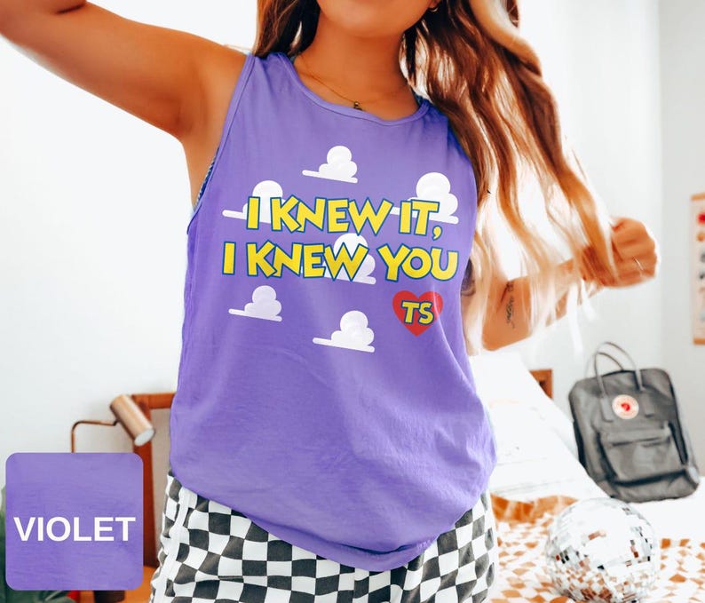 May include: A violet sleeveless tank top featuring a repeating white cloud pattern and yellow text that reads I KNEW IT, I KNEW YOU with a red heart containing the letters TS. The garment has a round neckline and a relaxed fit design.