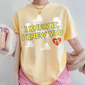 Może przedstawiać: Jasnoż&oacute;łty t-shirt z kr&oacute;tkim rękawem z niebieskim napisem I KNEW IT, I KNEW YOU, czerwonym sercem z ż&oacute;łtymi literami TS oraz grafikami białych chmur. Koszulka jest noszona z beżowym dzianinowym kardiganem i r&oacute;żowymi szortami w kratkę.