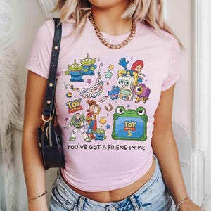 K&ouml;nnte beinhalten: Ein hellrosa T-Shirt mit kurzen &Auml;rmeln und einem farbenfrohen Grafikdruck verschiedener Toy Story-Figuren, darunter Woody, Buzz Lightyear und Aliens. Das Design enth&auml;lt den Text Disney Pixar, Toy Story, Toy Story 5 und You've Got A Friend In Me.