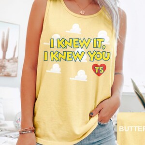 May include: A yellow tank top featuring the text I KNEW IT, I KNEW YOU in yellow with blue outlines, accompanied by white cloud graphics and a red heart containing the letters TS. The garment has a round neckline and sleeveless design.