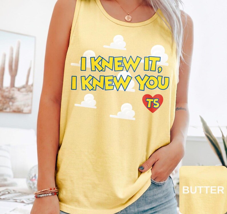 May include: A yellow tank top featuring the text I KNEW IT, I KNEW YOU in yellow with blue outlines, accompanied by white cloud graphics and a red heart containing the letters TS. The garment has a round neckline and sleeveless design.