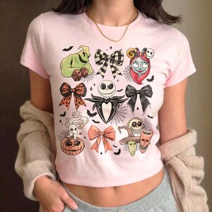 May include: A light pink t-shirt featuring various Halloween-themed character illustrations and bow designs. The graphics include Jack Skellington, Sally, Oogie Boogie, and other characters from The Nightmare Before Christmas, along with bats and spiderwebs.