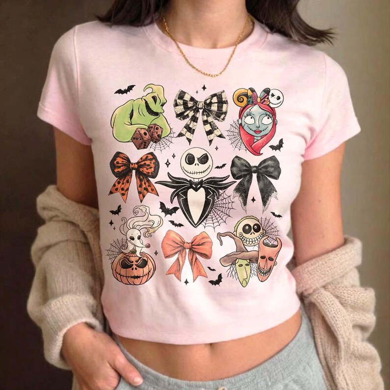 May include: A light pink t-shirt featuring various Halloween-themed character illustrations and bow designs. The graphics include Jack Skellington, Sally, Oogie Boogie, and other characters from The Nightmare Before Christmas, along with bats and spiderwebs.