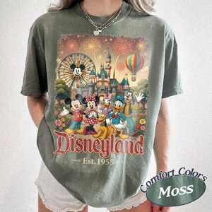 May include: A moss green t-shirt featuring a graphic print of Mickey Mouse, Minnie Mouse, Donald Duck, Daisy Duck, Goofy, and Pluto in front of a castle, a Ferris wheel, and a hot air balloon. The text reads Disneyland Est. 1955 in red lettering.