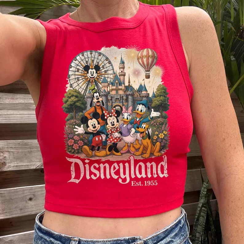 May include: A bright red sleeveless crop top featuring a graphic print of Mickey Mouse, Minnie Mouse, Goofy, Donald Duck, Daisy Duck, and Pluto in front of a castle, a Ferris wheel, and a hot air balloon. The text Disneyland Est. 1955 is printed below.