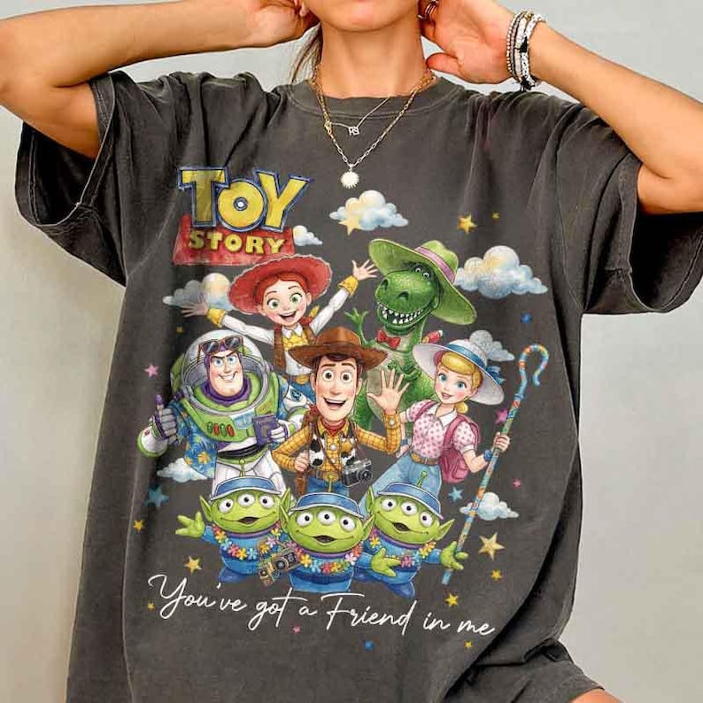 May include: Dark gray t-shirt featuring a colorful graphic of Toy Story characters, including Woody, Buzz Lightyear, and Rex. The shirt has the words "Toy Story" and "You've got a Friend in me" printed on it. The shirt is a crew neck.