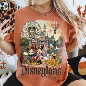 May include: A burnt orange crewneck t-shirt featuring a graphic print of Mickey Mouse, Minnie Mouse, Goofy, Donald Duck, Daisy Duck, and Pluto in front of a castle, a Ferris wheel, and a hot air balloon, with the text Disneyland Est. 1955.