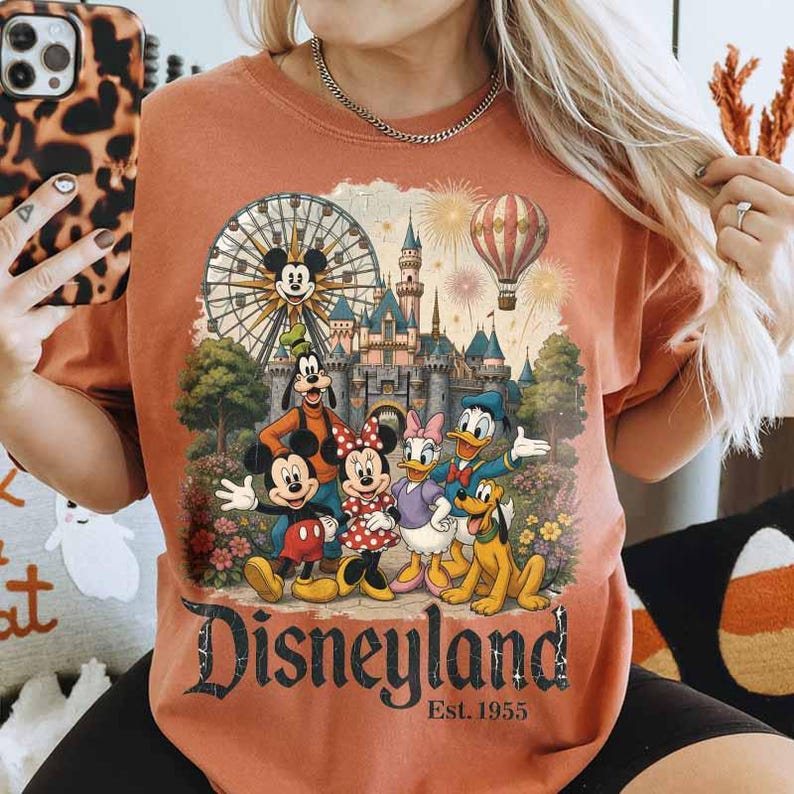 May include: A burnt orange crewneck t-shirt featuring a graphic print of Mickey Mouse, Minnie Mouse, Goofy, Donald Duck, Daisy Duck, and Pluto in front of a castle, a Ferris wheel, and a hot air balloon, with the text Disneyland Est. 1955.