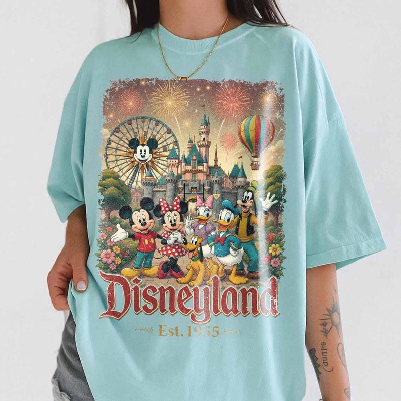 May include: A light blue t-shirt featuring a graphic print of Mickey Mouse, Minnie Mouse, Donald Duck, Daisy Duck, Goofy, and Pluto in front of a castle, a Ferris wheel, and fireworks. The text reads Disneyland Est. 1955 in a red vintage font.