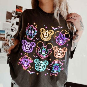 May include: Dark gray t-shirt featuring a colorful neon design of Disney characters in Halloween costumes. The graphic includes Donald Duck, Mickey Mouse, Minnie Mouse, and other characters with bats and stars.