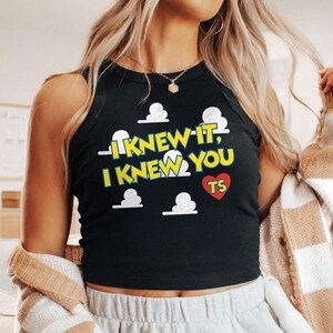 May include: A black sleeveless tank top featuring a graphic print of white clouds and the yellow text I KNEW IT, I KNEW YOU with a red heart containing the letters TS. The garment has a high neckline and a ribbed collar design.