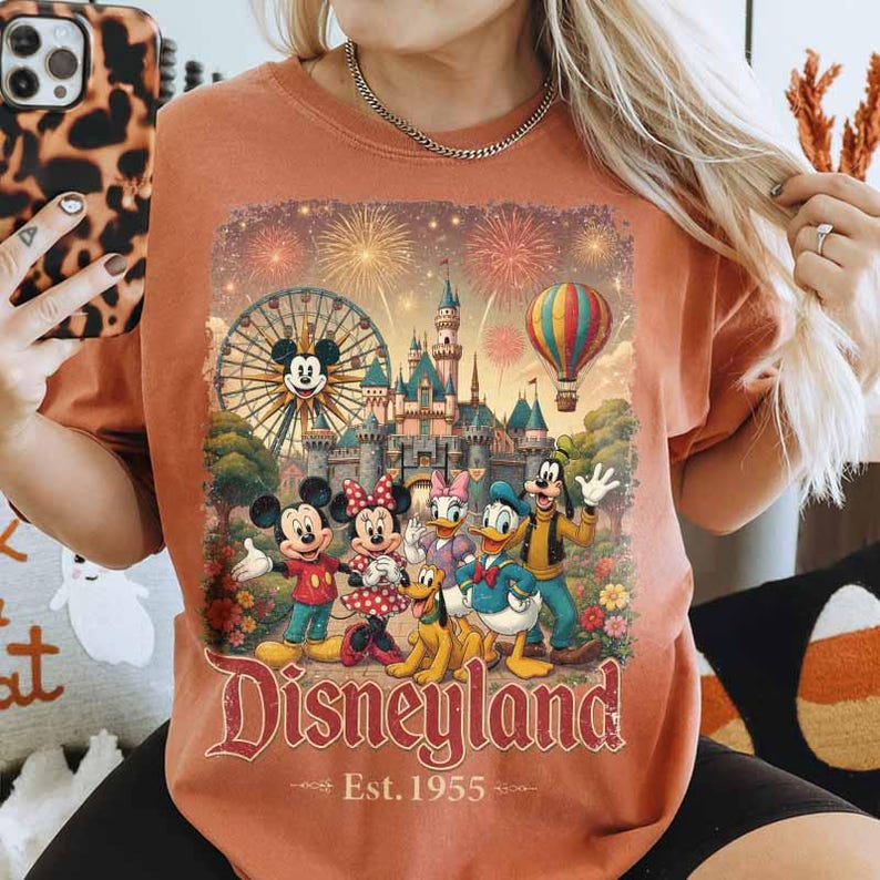 May include: A rust-colored t-shirt featuring a graphic print of Mickey Mouse, Minnie Mouse, Donald Duck, Goofy, and Pluto in front of a castle, a Ferris wheel, and a hot air balloon with fireworks. The text reads Disneyland Est. 1955.