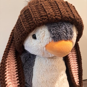 May include: A plush penguin toy wearing a brown knit hat with ear flaps. The penguin has a white body, gray wings, and an orange beak. The hat has a brown pom-pom on top. The ear flaps are lined with pink.