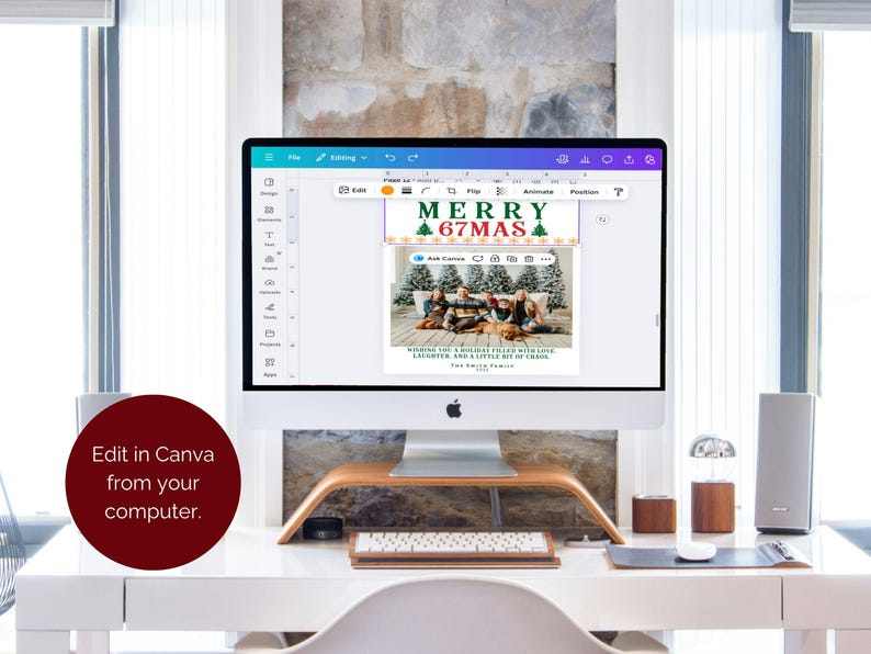 May include: A computer monitor displays a Christmas card design with the text "MERRY 67MAS" and a family photo. The card is being edited in Canva. The computer sits on a white desk with a wooden stand, keyboard, and speakers.