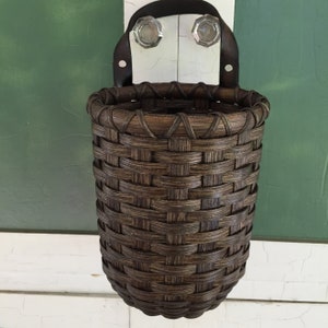 May include: A dark brown woven basket with a leather strap and metal hooks for hanging. The basket is empty and ready to be filled with items.