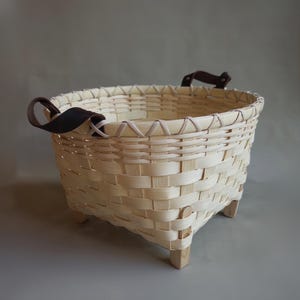 May include: A round woven basket made of light-colored wood strips featuring a reinforced rim, four small wooden feet at the base, and two dark brown leather handles attached with metal rivets on opposite sides of the upper edge.