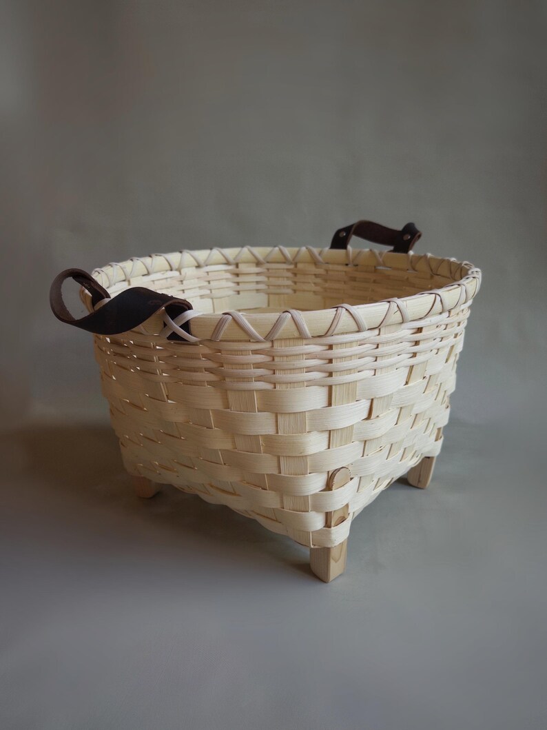 May include: A round woven basket made of light-colored wood strips featuring a reinforced rim, four small wooden feet at the base, and two dark brown leather handles attached with metal rivets on opposite sides of the upper edge.