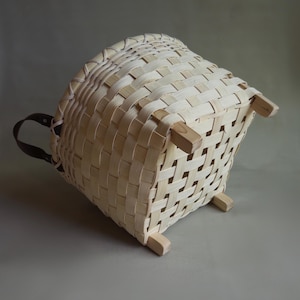 May include: A light-colored woven basket with a square base and four wooden feet. The basket features a grid-like pattern of thin, flat strips and includes a dark brown leather handle attached to one side, providing a contrast to the light wood.