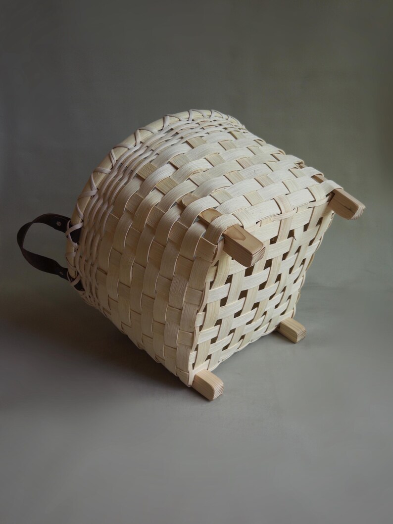 May include: A light-colored woven basket with a square base and four wooden feet. The basket features a grid-like pattern of thin, flat strips and includes a dark brown leather handle attached to one side, providing a contrast to the light wood.