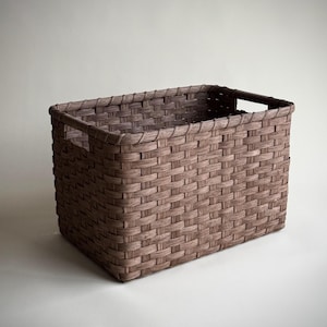 May include: A rectangular woven basket in a warm brown hue. The basket features a tightly woven pattern with integrated handles on the short sides. The neutral color and simple design suggest its use for storage or organization.