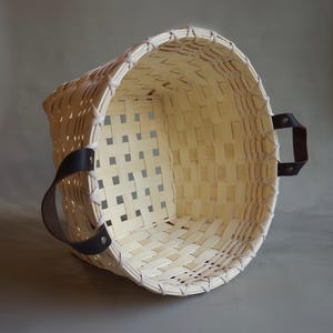 May include: A round, woven basket made of light-colored, natural wood strips featuring a wide, open top and a grid-like pattern. The basket includes two dark brown leather handles securely attached to the sides with silver-colored metal rivets.
