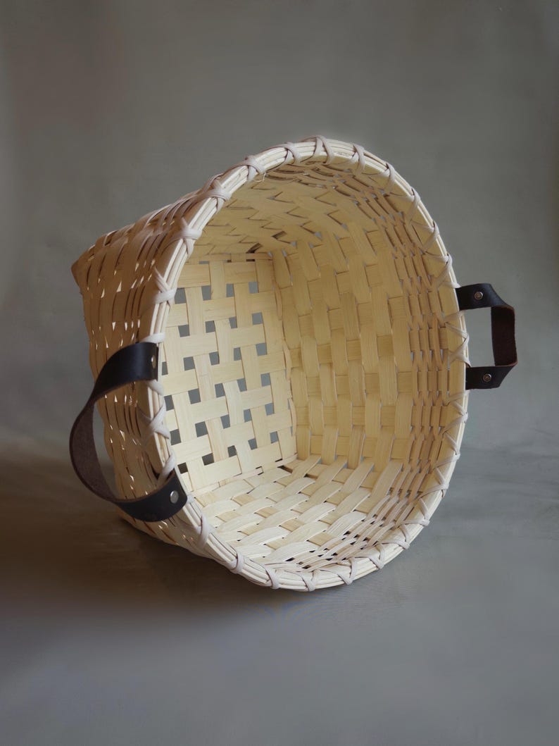 May include: A round, woven basket made of light-colored, natural wood strips featuring a wide, open top and a grid-like pattern. The basket includes two dark brown leather handles securely attached to the sides with silver-colored metal rivets.