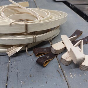 May include: A coiled bundle of light-colored, flexible wooden strips tied with twine, accompanied by several small, light-colored wooden blocks and dark brown textured leather loops secured with silver-toned metal rivets.