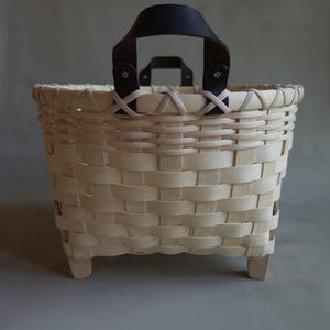 May include: A woven light-colored wood basket featuring a square base with four small feet. The basket has a wide, open top rim and two dark brown leather handles attached with metal rivets to the top edge of the woven structure.