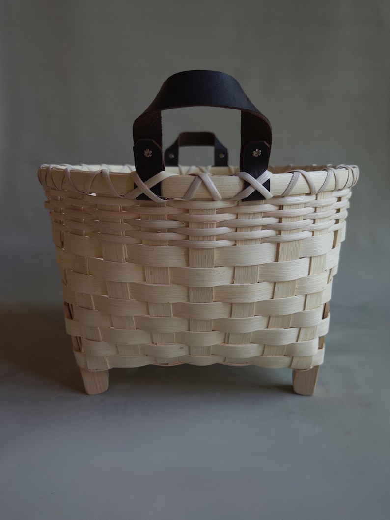 May include: A woven light-colored wood basket featuring a square base with four small feet. The basket has a wide, open top rim and two dark brown leather handles attached with metal rivets to the top edge of the woven structure.